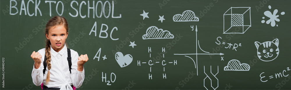 horizontal image of angry schoolgirl showing clenched fists near chalkboard with back to school lettering and geometry formulas in classroom