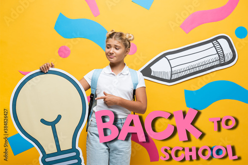 Confused schoolgirl pointing with finger at paper light bulb near decorative elements and back to school lettering on yellow