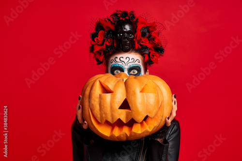 Halloween Party girl. Happy girl with sugar skull makeup, with a wreath of flowers on her head and skull, wearth lace gloves and leather jacket, Holds a big Jack-o-lantern pumpkin isolated on red photo