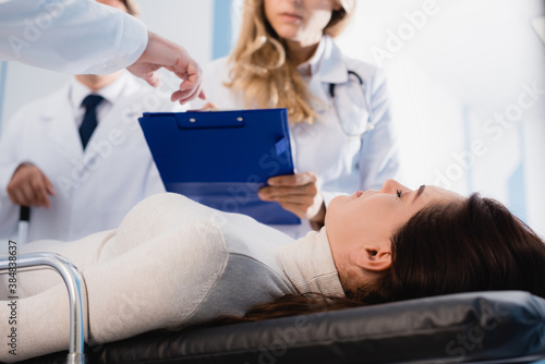 Selective focus of patient with closed eyes lying near doctor pointing at clipboard to colleague in clinic