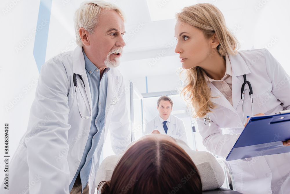Selective focus of senior doctor talking to colleague with clipboard near patient on stretcher in clinic Selective focus of senior doctor talking to colleague with clipboard near patient on stretcher in clinic