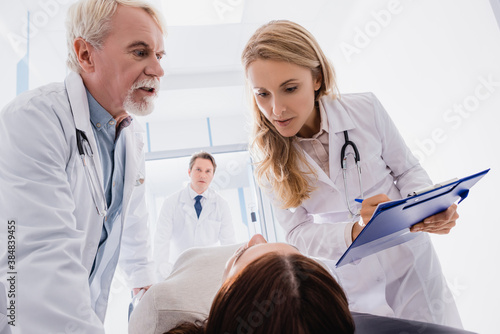 Selective focus of doctor writing on clipboard near colleague and ill patient in clinic
