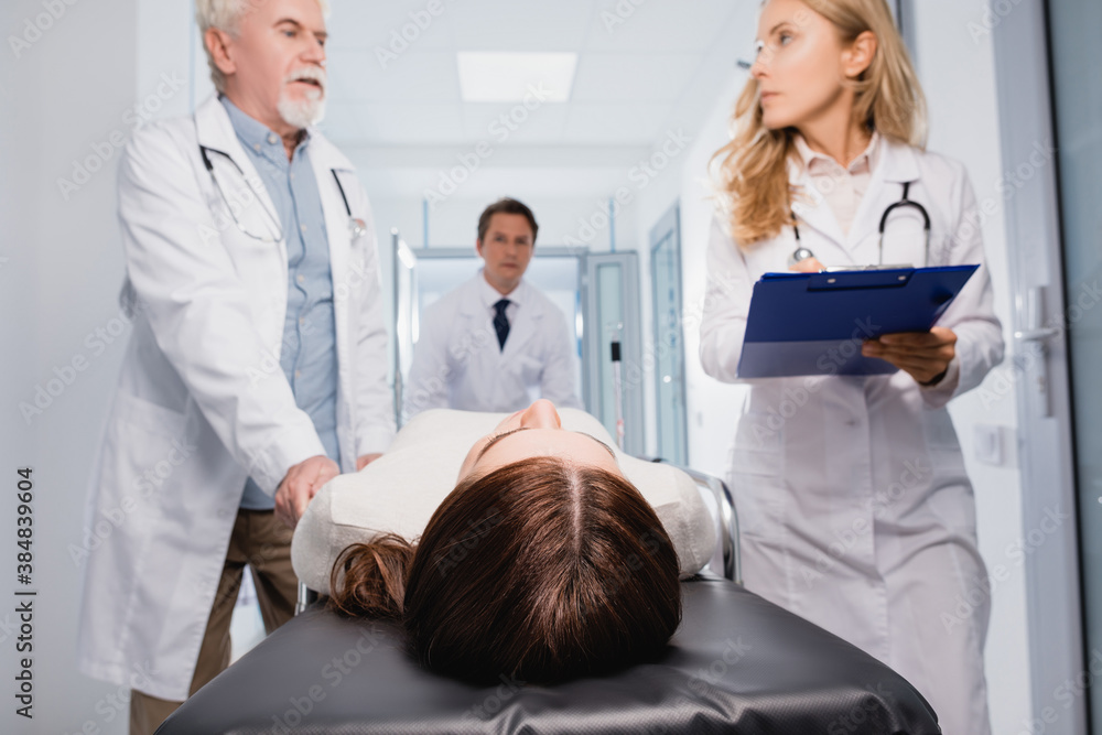 Selective focus of sick woman lying on stretcher near doctors with clipboard in clinic