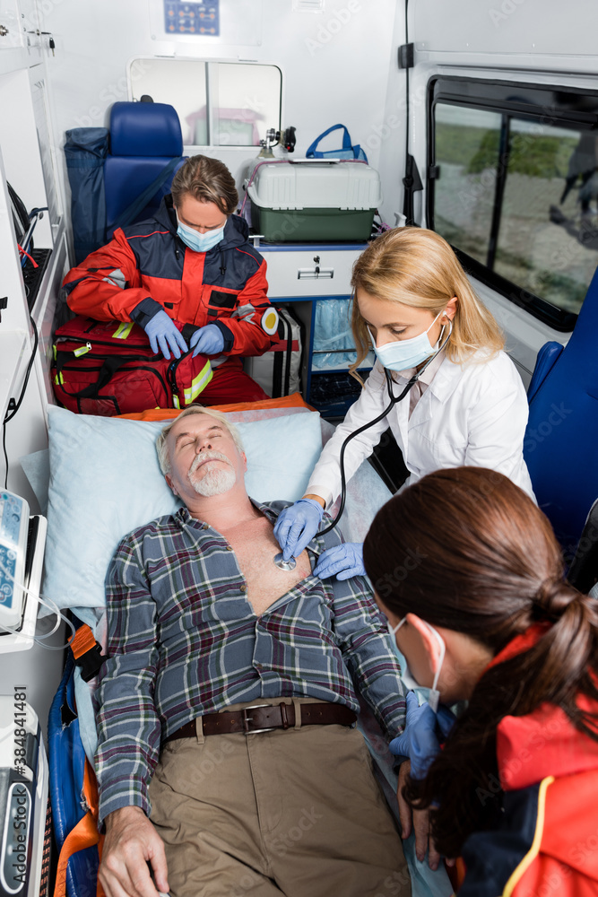 Selective focus of doctor in medical mask examining patient with stethoscope near paramedics in ambulance car Selective focus of doctor in medical mask examining patient with stethoscope near paramedics in ambulance car