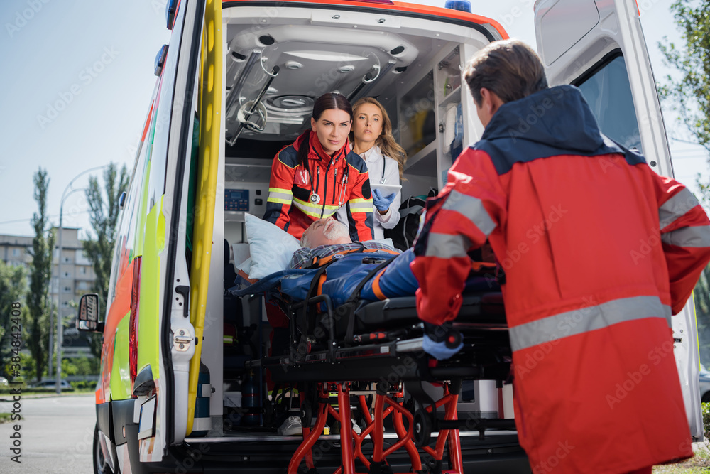 Selective focus of doctor with digital tablet standing in ambulance car near paramedics with patient on stretcher outdoors Selective focus of doctor with digital tablet standing in ambulance car near paramedics with patient on stretcher outdoors