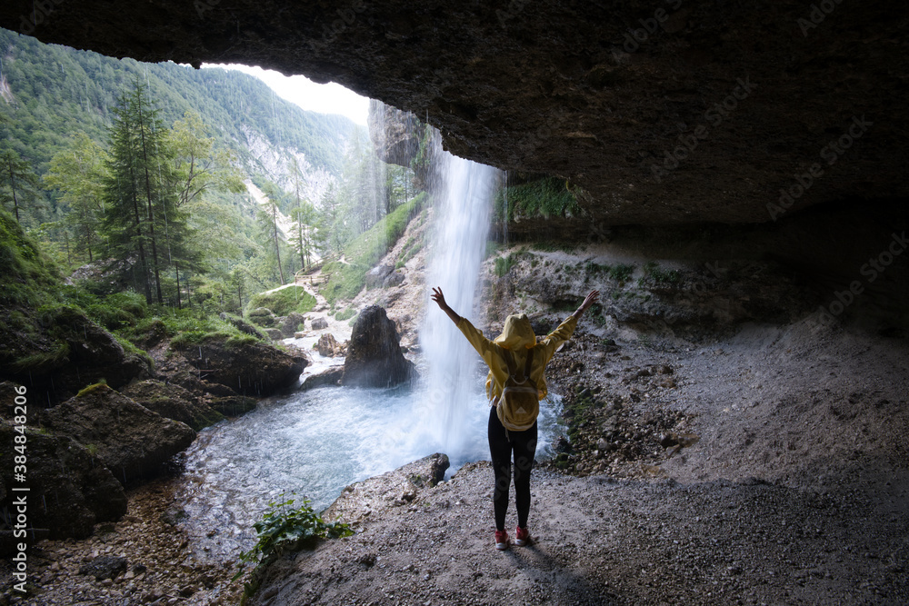 Obraz premium Young woman standing in front of waterfall with her hands outstretched. Caucasian female tourist in forest with her arms wide open