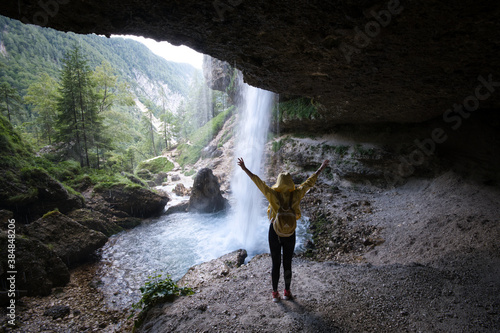 Young woman standing in front of waterfall with her hands outstretched. Caucasian female tourist in forest with her arms wide open