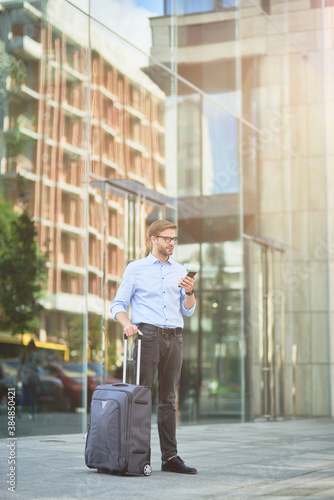 Vertical shot of elegant handsome businessman with suitcase using smartphone and looking away while standing outdoors, waiting for taxi