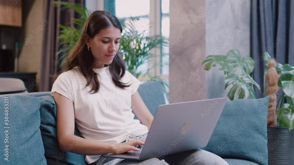 Cute young woman sitting on couch using laptop notebook typing message. Social media studying or working online at home. Slow motion Cute young woman sitting on couch using laptop notebook typing message. Social media studying or working online at home. Slow motion