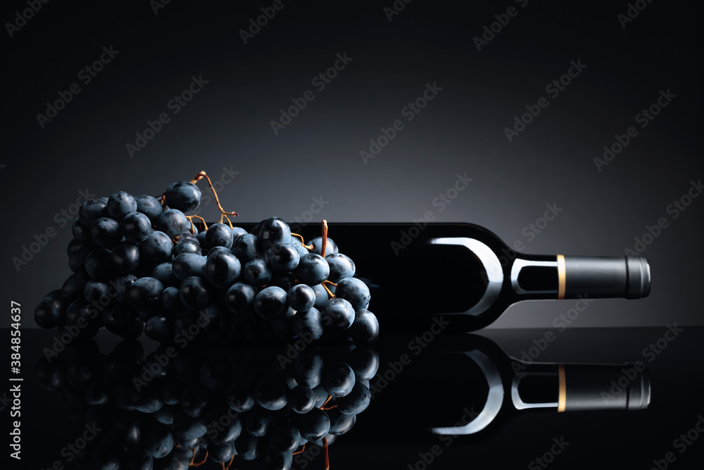 Bottle of red wine and a bunch of grapes on a black reflective background. - obrazy, fototapety, plakaty 