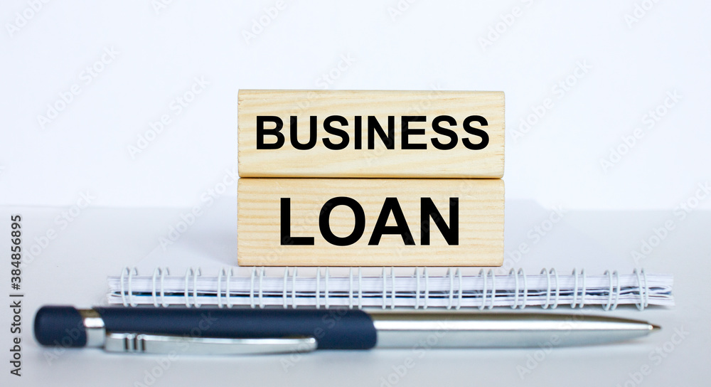 The text Business Loan on a bar of jenga wood, lying on a Notepad with a metal blue pen The text Business Loan on a bar of jenga wood, lying on a Notepad with a metal blue pen