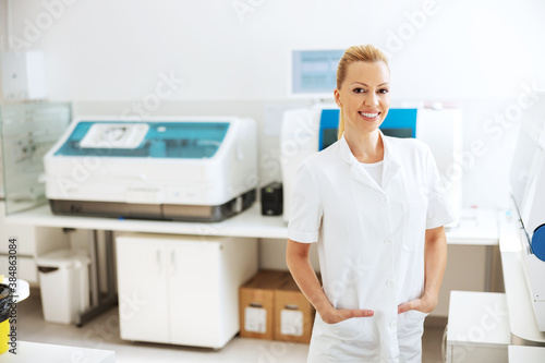 Smiling attractive blond lab assistant in white sterile uniform standing in laboratory with hands in pockets and looking at camera.