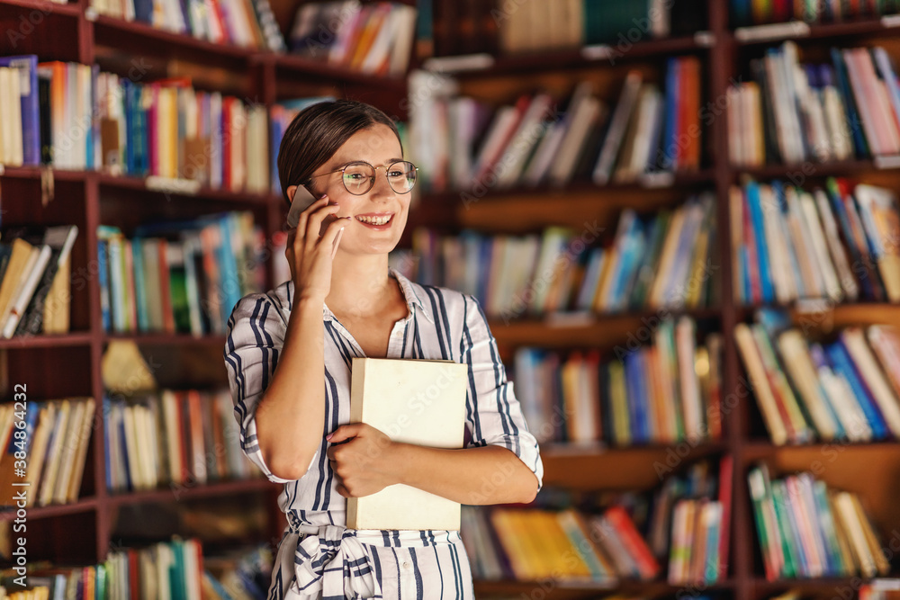 Young smiling attractive college girl with eyeglasses and in dress standing in library, holding book and having phone call. Young smiling attractive college girl with eyeglasses and in dress standing in library, holding book and having phone call.