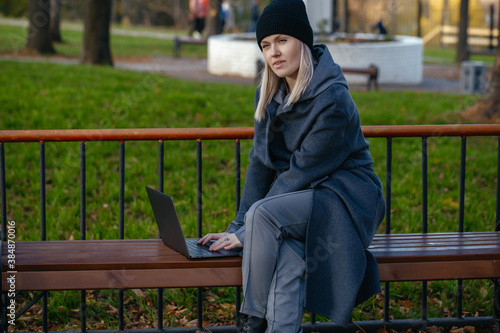 beautiful girl in a coat and hat is sitting at a laptop and doing work in the Park. Outdoor activities.