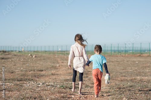 two poor children family brother with toy and thin sister refugee illegal immigrant walking barefooted through hot desert towards state border with barbed fence wire