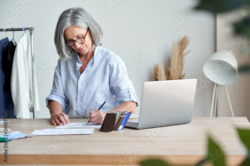 Middle aged stylish woman fashion designer drawing sketches in studio. Mature old adult elegant grey-haired lady dressmaker small business owner creating new fashion design cloth in atelier.