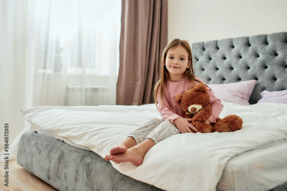 A Cute Little Browneyed Girl Sitting Barefoot On A Huge Bed Holding Her Teddybear With Her Feet