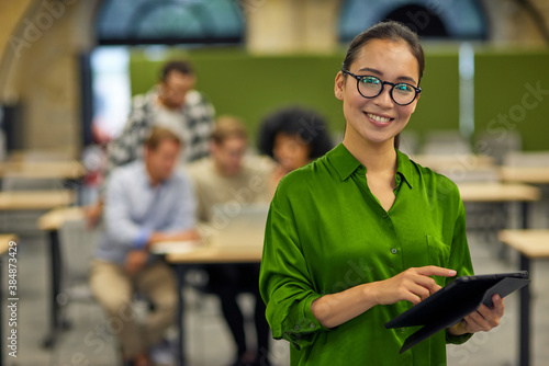 Portrait of young happy asian woman using digital tablet, looking at camera and smiling while standing in the coworking space with colleagues on the background