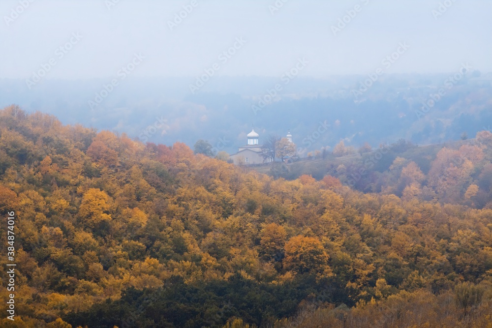 mountains covered with magnificent mountain forest, green, yellow and orange autumn leaves, cloudy and misty October morning, Orthodox church in poor visibility, ecotourism active rest concept