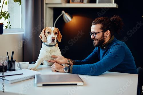 Young smiling attractive hipster sitting in his home office and playing with his beloved pet.
