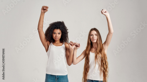Portrait of two young diverse women, best friends looking serious at camera while raising arm and making a pinkie promise sign isolated over grey background