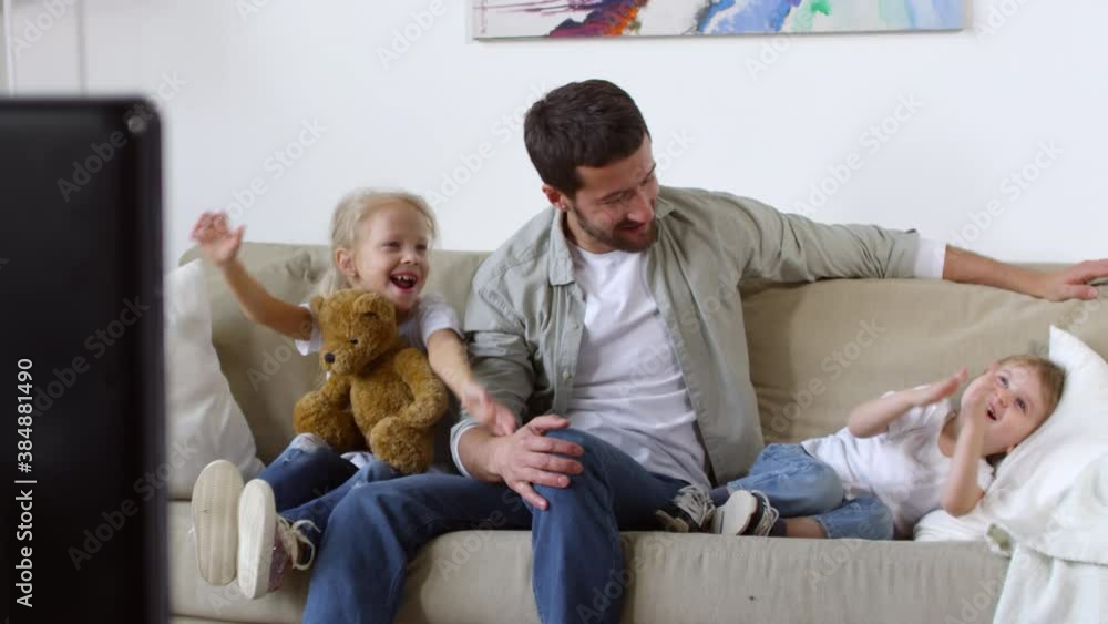 Dolly-in shot of beautiful woman walking into living room with bowl of candies and sitting down on sofa to watch TV with excited little kids and happy husband
