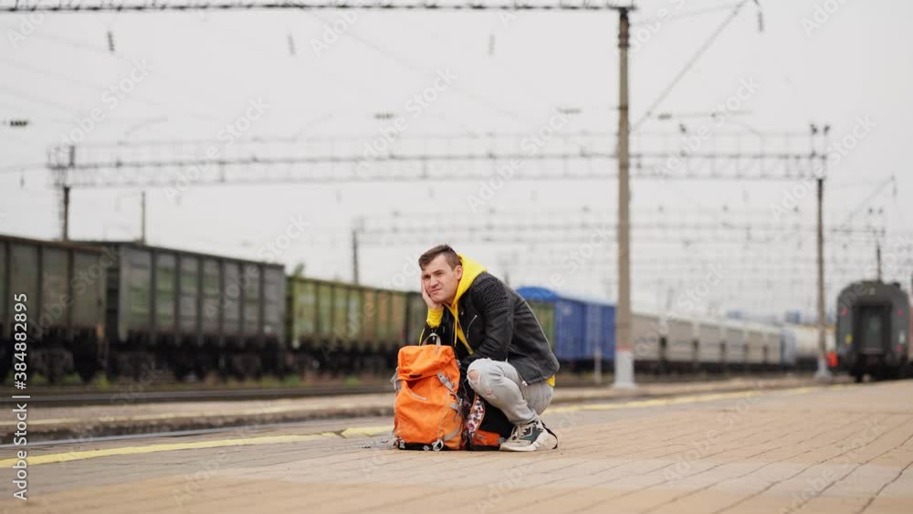 Young man squats on platform, waiting for train. Male passenger with backpacks sitting on railroad platform in waiting for train ride. Concept of tourism, travel and recreation. Young man squats on platform, waiting for train. Male passenger with backpacks sitting on railroad platform in waiting for train ride. Concept of tourism, travel and recreation.