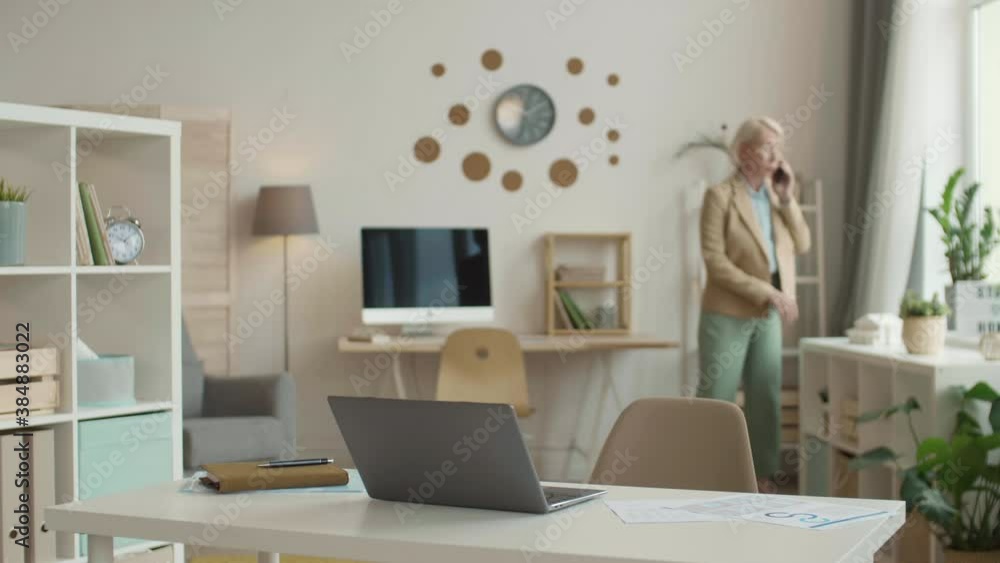 Locked-down of woman wearing modern formal suit walking into home office talking on phone, hanging up, sitting down on chair at desk, starting video call on laptop Locked-down of woman wearing modern formal suit walking into home office talking on phone, hanging up, sitting down on chair at desk, starting video call on laptop