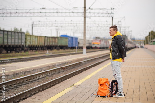 Young man stands on platform, waiting for train. Male passenger with backpacks on railroad platform in waiting for train ride. Concept of tourism, travel and recreation.
