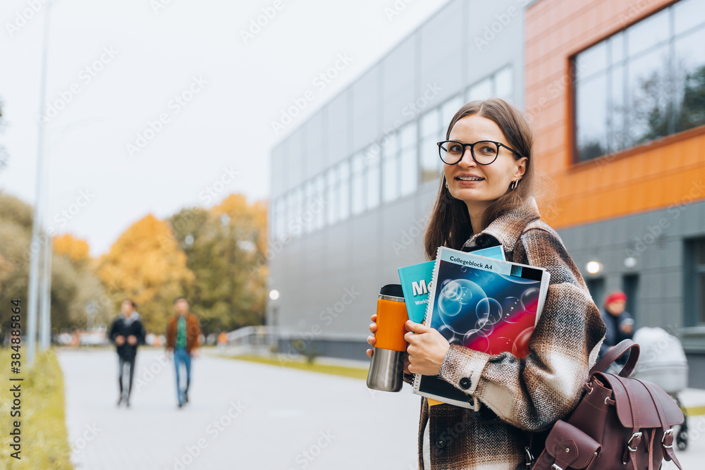 Fototapeta premium Portrait of girl student standing at university campus. Laughing Woman wearing braces, eyeglasses, bag with books having coffee break after lecture. Enjoying College life. Learning education concept.