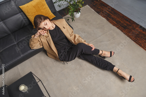Stylishly dressed young caucasian woman in beige blazer and black pants sitting on the floor at sofa. Interior fashion portrait