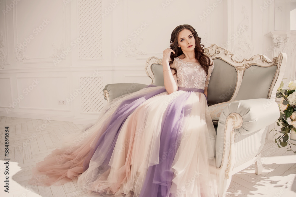 Curly girl in an original dress sits on a sofa. Beautiful model in a beautiful wedding dress in a white photo studio. Bride in color wedding dress. Curly girl in an original dress sits on a sofa. Beautiful model in a beautiful wedding dress in a white photo studio. Bride in color wedding dress.