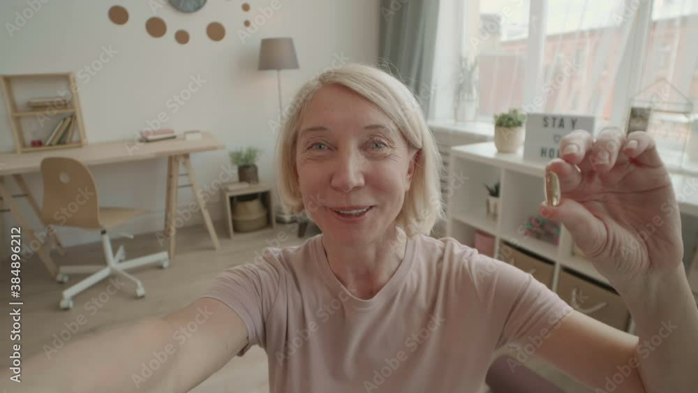 POV portrait of lively fit senior female vlogger using selfie camera to film for video blog holding transparent supplement pill smiling and talking on camera in living room POV portrait of lively fit senior female vlogger using selfie camera to film for video blog holding transparent supplement pill smiling and talking on camera in living room