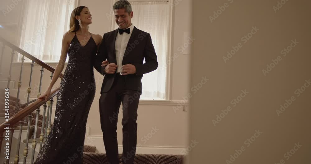 Attractive couple talking while moving down the stairs in a mansion. Woman in evening gown and man in tuxedo walking down the steps hand in hand.