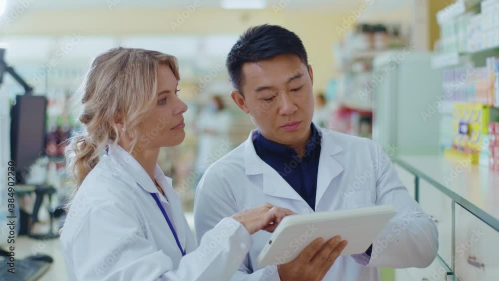 Close up woman and Asian man cooperating in drugstore use tablet computer. Attracitve woman apothecarist talking with male colleague learning pills. Slow motion Close up woman and Asian man cooperating in drugstore use tablet computer. Attracitve woman apothecarist talking with male colleague learning pills. Slow motion