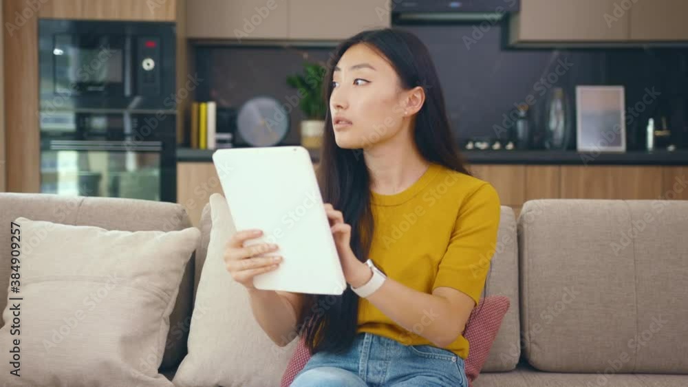 Close up young asian woman sitting on couch in cozy room use tablet device smile at home. Businesswoman internet confident chatting. Portrait. Slow motion Close up young asian woman sitting on couch in cozy room use tablet device smile at home. Businesswoman internet confident chatting. Portrait. Slow motion