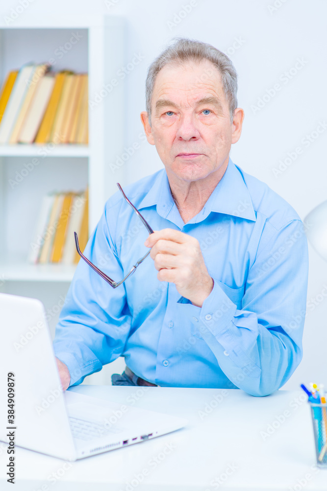 A male pensioner with glasses sits behind a laptop screen in his office. Work for seniors concept