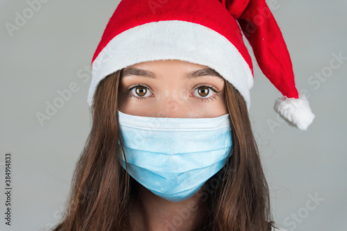 A young woman in a Santa Claus hat and a medical mask looks into the camera. Celebrating the new year in a pandemic. . High quality photo