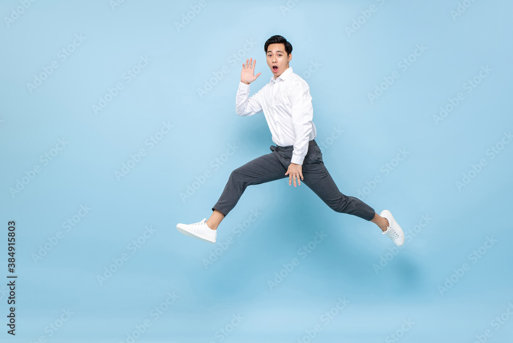 Fun energetic Asian man in semi formal clothes jumping in mid air isolated on light blue background