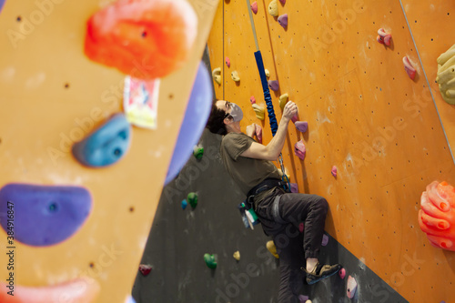 Man wearing COVID-19 or coronavirus pandemic mask climbs a wall attached to an auto belay rope in a climbing gym while following social distance guidelines
