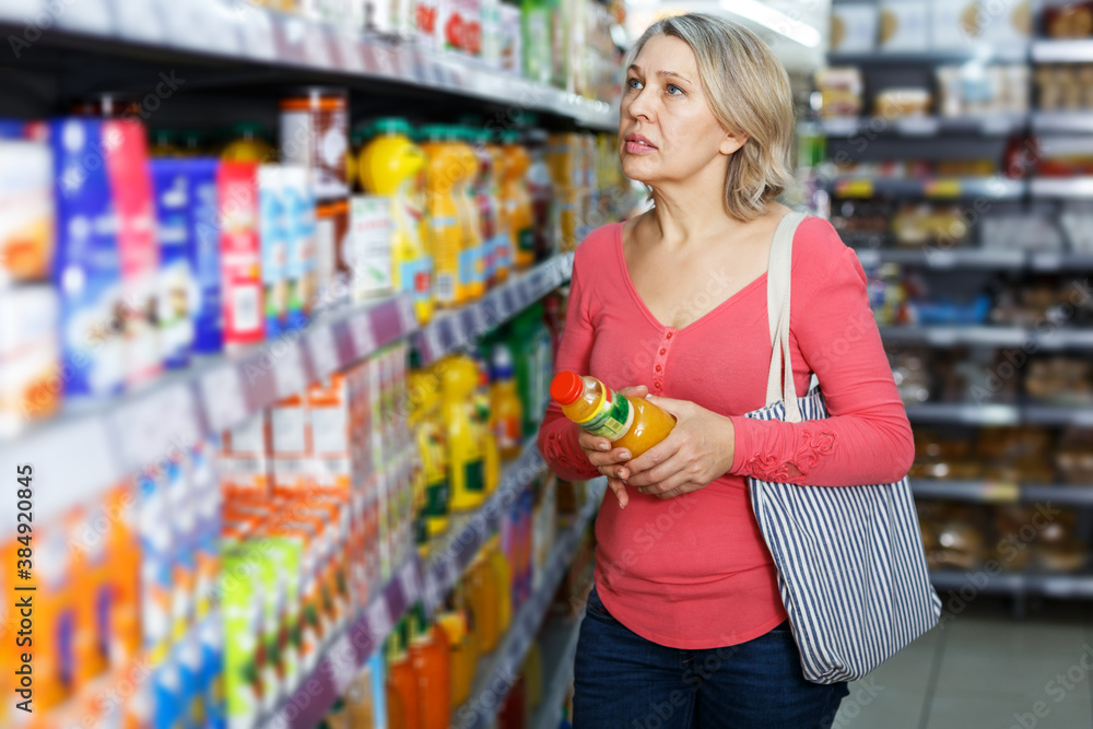 Middle aged blonde woman choosing and buying refreshing fruit drinks at grocery store