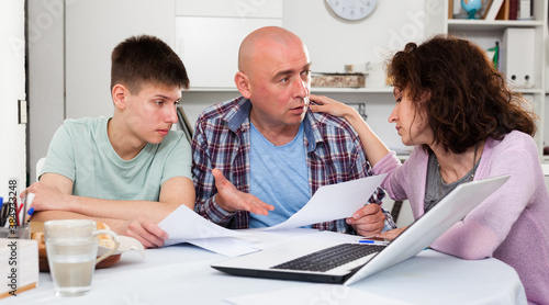 Chagrined man sitting at home table with papers while family soothing him..