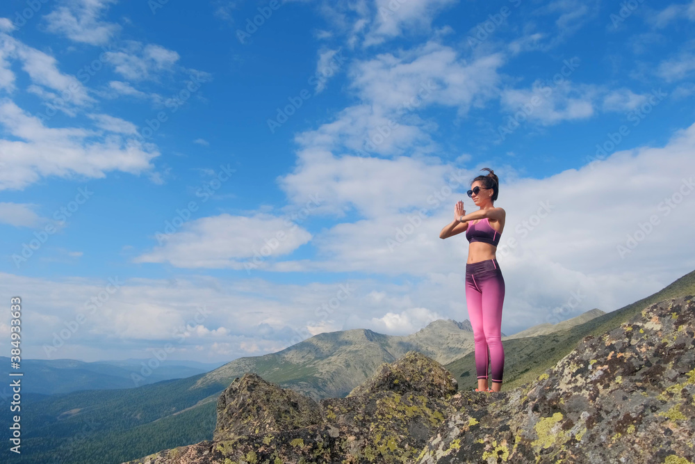 Naklejka premium A woman doing yoga exercises in prayer posture on top of mountains. Young woman hiking up hill against a blue sky with clouds.
