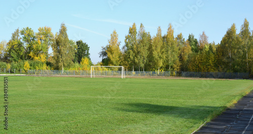 In an empty stadium, a green football field and white gates against the backdrop of an autumn forest and blue sky.