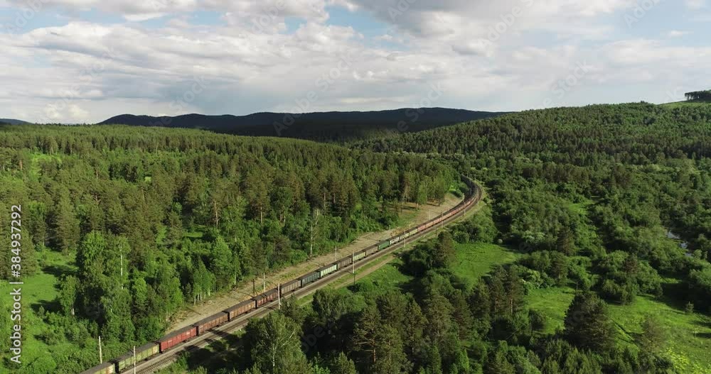 Freight long train carries with coal carriages an electric locomotive by dangerous part of two-sided Trans Siberian railways in forest mountains / Aerial drone wide view at summer sunny day