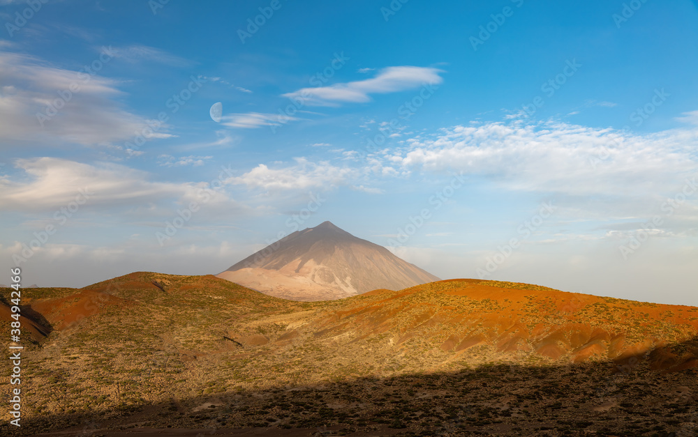 A panoramic view of the Teide region in the early morning after sunrise with the Pico del Teide in the center. The sun is shining in a blue sky with beautiful clouds.