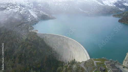 Arch dam and reservoir lake in Swiss Alps mountains feeding hydropower electric power plant to produce renewable energy, sustainable hydroelectricity, snowy weather, aerial drone footage 4K 60 fps