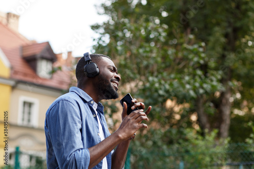 Walking in the street, relaxation, leisure. Young Afro-American man in headphones listening music on smart phone using music app. Portrait of smiling guy in earphones and mobile phone outdoors.