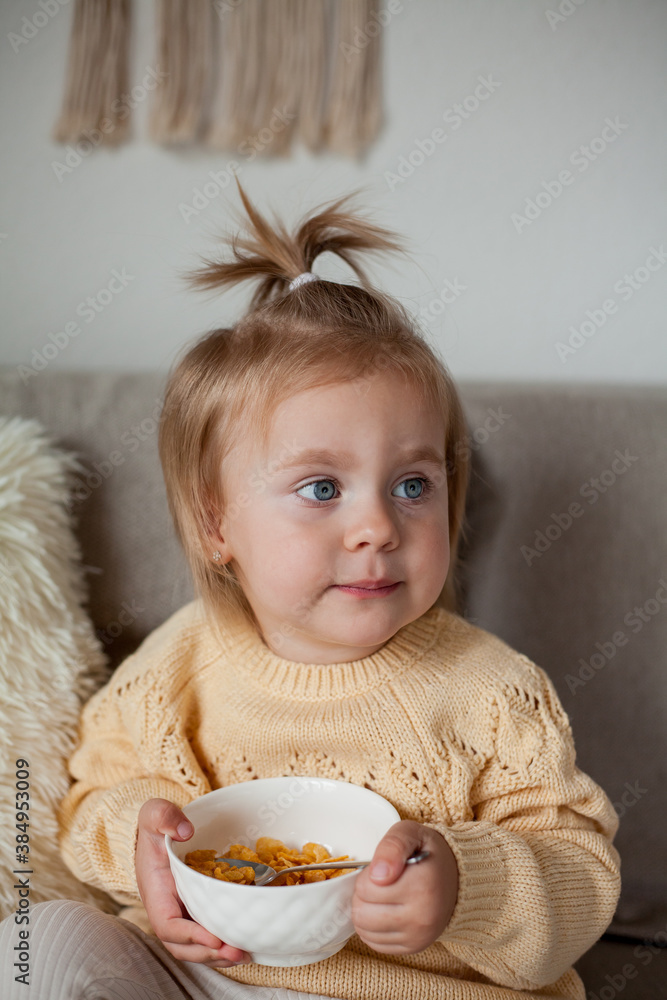 A cute little girl 2 years old in a cozy knitted sweater is having breakfast on the couch. Cozy. Fall. A cute little girl 2 years old in a cozy knitted sweater is having breakfast on the couch. Cozy. Fall.