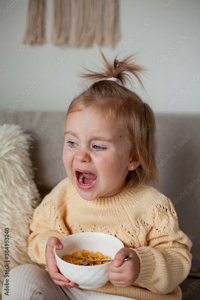 A cute little girl 2 years old in a cozy knitted sweater is having breakfast on the couch. Cozy. Fall.